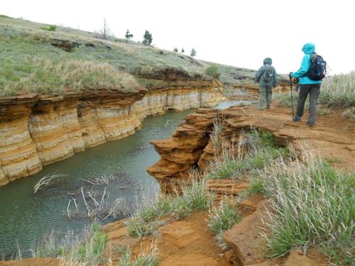 Two hikers in rain jackets stand by a river, surrounded by layered rock formations and grassy terrain.
