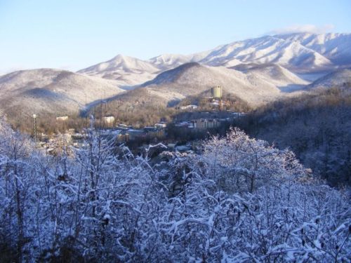 Snow-covered mountains and a valley with buildings, under a clear blue sky. Winter landscape with trees in the foreground.