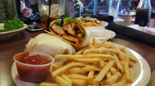 A plate with fries, a grilled chicken wrap, and a small cup of salsa, with drinks and other dishes in the background.