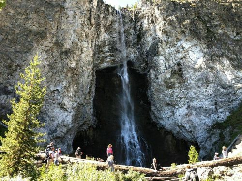 A waterfall cascades down rocky cliffs, surrounded by trees and people exploring the area below.