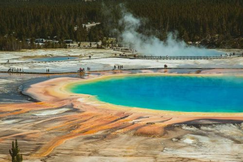 A vibrant geothermal pool with blue and orange hues, surrounded by steam and visitors on a wooden walkway in a natural landscape.