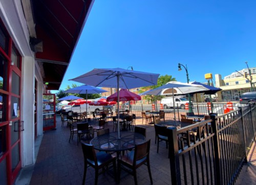 Outdoor dining area with tables and umbrellas, surrounded by a black fence under a clear blue sky.