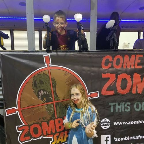 Two children with face paint stand behind a banner for a zombie-themed event, holding toy guns and smiling.