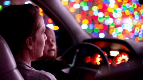 A woman and child inside a car, looking at colorful holiday lights outside.