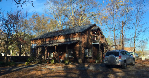 A rustic wooden building surrounded by trees, with a car parked nearby and a clear blue sky above.