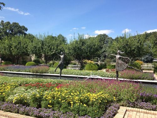 Two dancing sculptures in a vibrant garden filled with colorful flowers and lush greenery under a clear blue sky.