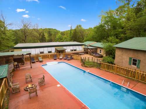Aerial view of a pool surrounded by wooden decking, chairs, and green trees under a clear blue sky.