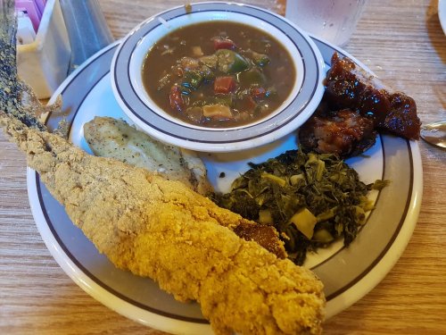 A plate featuring fried fish, collard greens, a bowl of gumbo, and a side of cornbread.