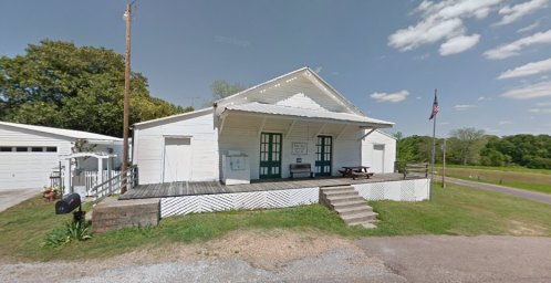A white wooden building with green doors, a porch, and an American flag, set in a rural area under a blue sky.