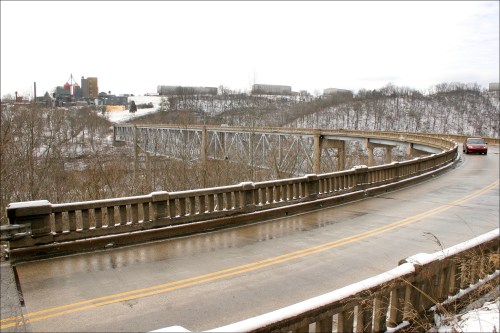 A curved bridge spans a snowy landscape, with a car driving along the road and industrial buildings in the background.