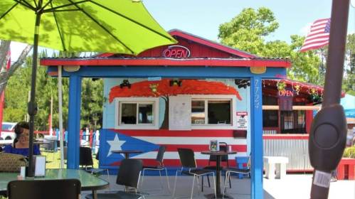 Colorful food stand with a Puerto Rican flag, outdoor seating, and an "Open" sign on a sunny day.