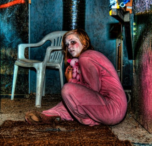 A girl in a pink onesie sits on the floor, holding a stuffed animal, with a distressed expression in a dimly lit room.