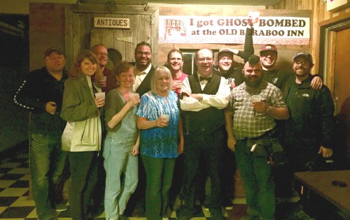 A group of people poses together at a bar, smiling and holding drinks, with a sign in the background about ghost bombing.