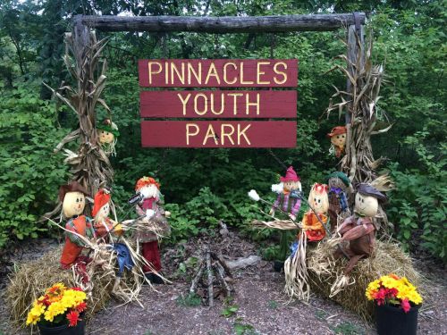 A colorful sign reading "Pinnacles Youth Park" surrounded by scarecrows and hay bales in a green, wooded area.