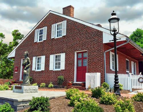 Historic brick house with white shutters, a red door, and a statue in front, surrounded by greenery and a lamp post.