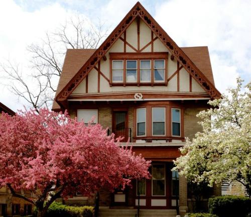 A charming two-story house with a steep roof, surrounded by blooming pink and white trees under a cloudy sky.