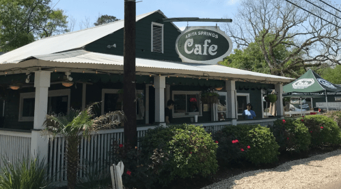 A green café with a sign reading "Abita Springs Café," surrounded by plants and flowers on a sunny day.