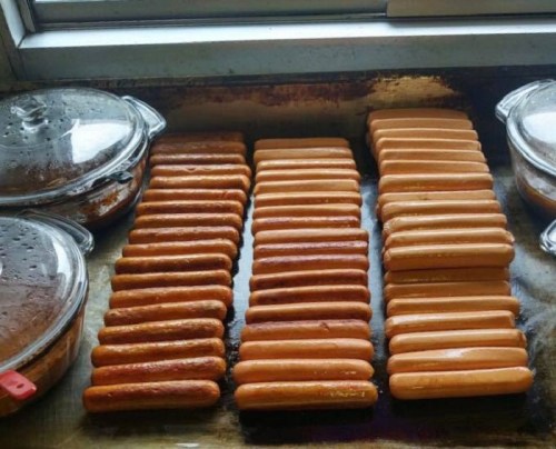 A tray of cooked hot dogs arranged in neat rows, with glass containers in the background.