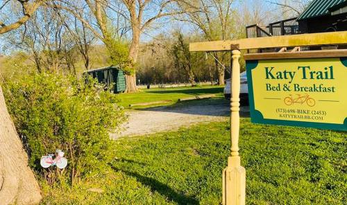 Sign for Katy Trail Bed & Breakfast with a green background, surrounded by trees and a gravel path.