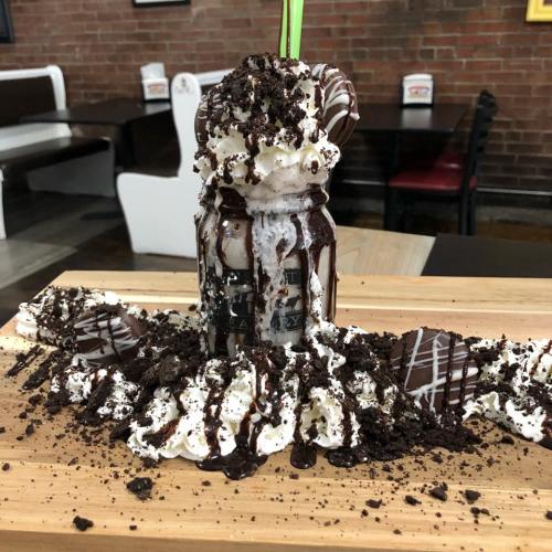 A tall milkshake topped with whipped cream, chocolate drizzle, and crushed cookies, served on a wooden platter.