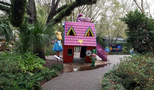 Colorful playhouse with a pink roof, surrounded by greenery, featuring whimsical characters and a winding path.