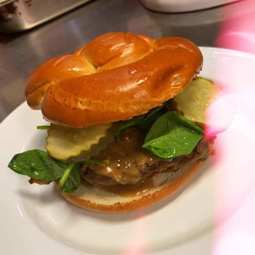 A close-up of a burger with a shiny bun, spinach, pickles, and a savory sauce on a white plate.
