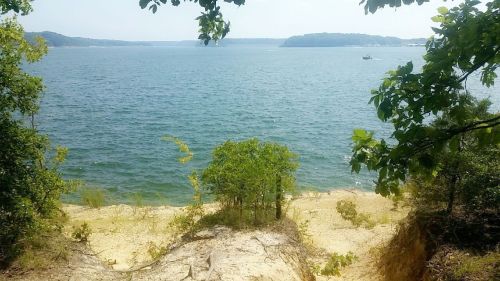 A serene lakeside view with sandy shores, surrounded by trees and distant hills under a clear blue sky.