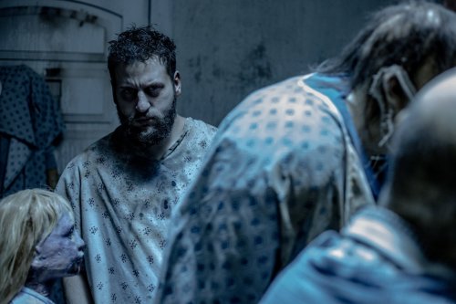 A dimly lit room with a man in a hospital gown, surrounded by eerie, unsettling figures and a doll.