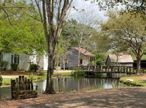 A peaceful scene featuring a pond, wooden bridge, and historic buildings surrounded by trees and greenery.