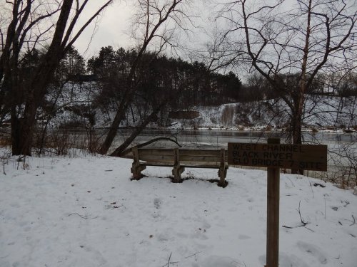 A snowy riverside scene featuring a wooden bench and a sign for the West Channel of the Black River.
