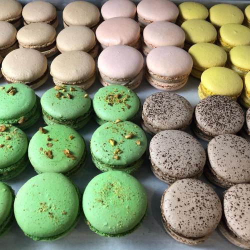 A variety of colorful macarons arranged in rows, featuring green, pink, yellow, and brown flavors with toppings.