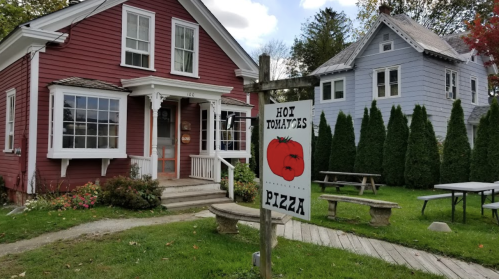 A red house with a sign for "Hot Tomatoes Pizza" in front, surrounded by green grass and trees.