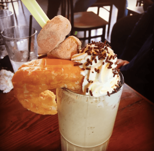 A thick milkshake topped with whipped cream, sprinkles, a donut, and a shrimp-shaped garnish on a straw.