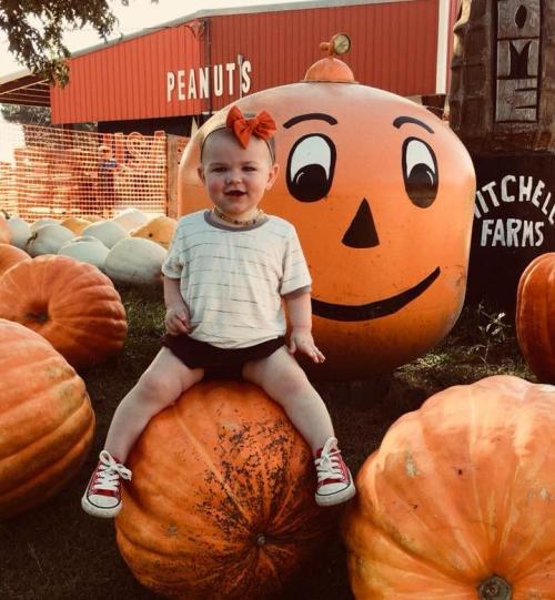 A smiling toddler with a bow sits on a large pumpkin, with a cheerful pumpkin decoration in the background.