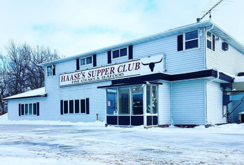 Exterior of Haase's Supper Club, a white building with a sign for fine steaks and seafood, surrounded by snow.