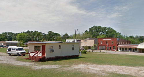 A small white building with a porch in a grassy area, surrounded by a few other buildings and trees in the background.