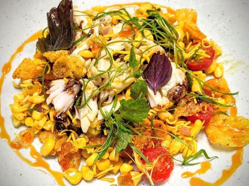 A colorful dish featuring crab, corn, cherry tomatoes, and crispy elements, garnished with fresh herbs on a white plate.