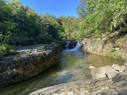 A serene natural scene featuring a small waterfall surrounded by lush greenery and rocky terrain under a clear blue sky.