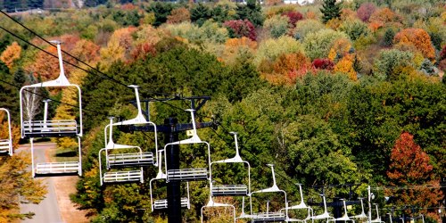 A ski lift with empty chairs overlooks a vibrant forest of autumn-colored trees.