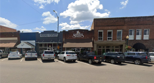 A street view of a small town with shops and parked cars under a blue sky with fluffy clouds.
