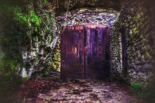 A weathered metal door set in a rocky, overgrown wall, surrounded by greenery and stone pathways.