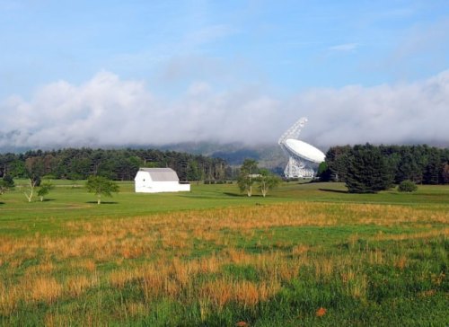 A white barn stands in a green field with a large satellite dish and trees under a cloudy blue sky.