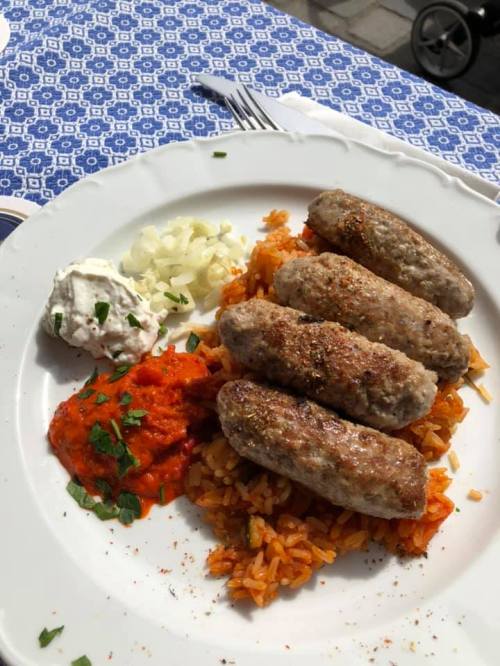 A plate of four grilled sausages on a bed of rice, accompanied by tomato sauce, yogurt, and chopped onions.
