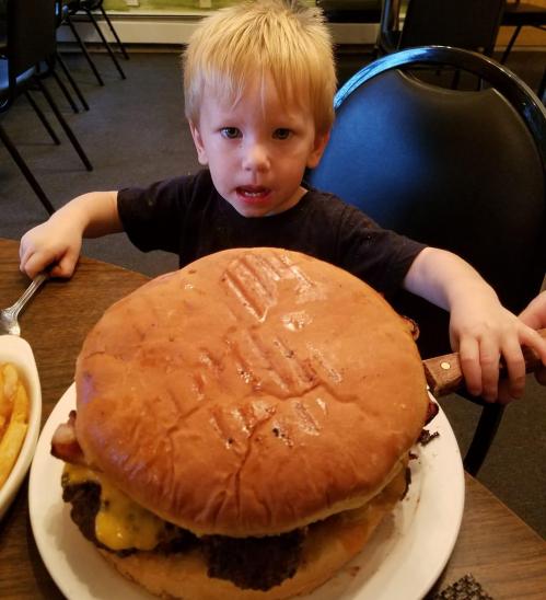 A young child sits at a table, holding a giant burger that's larger than their head, with fries in the background.