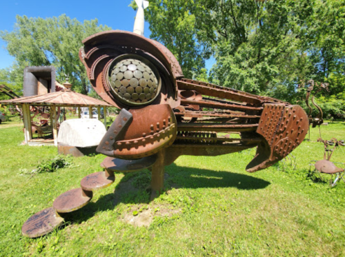 A large, abstract metal sculpture resembling a bird, set in a green outdoor space with trees in the background.