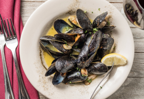 A plate of mussels in garlic butter, garnished with parsley and a lemon wedge, served with silverware and a glass of wine.