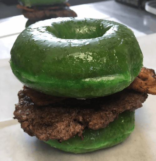 A green glazed donut sandwiching a beef patty and crispy bacon, resting on a white surface.