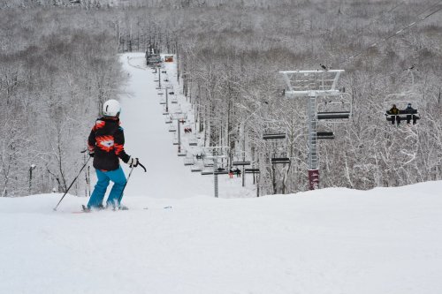 A skier in colorful gear stands on a snowy slope, looking down at a ski lift and tree-covered hills in the background.