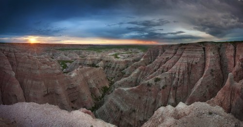 A panoramic view of rugged, colorful canyons at sunset, with dramatic clouds in the sky above.