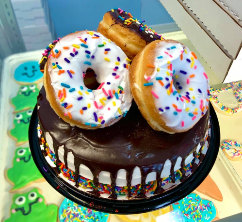 A chocolate cake topped with two glazed donuts and colorful sprinkles, displayed on a vibrant background.